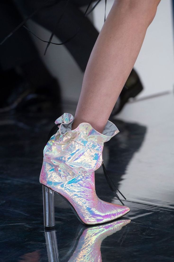 loseup image of a woman's leg showcasing iridescent, highheeled ankle boots. the boots have a ruffled, crumpled effect and are made of a shimmering, holographic material with a pearlescent sheen, reflecting light in various colors.  the heels are clear and cylindrical. the model's leg is slender, and the background is blurred but shows a runway or similar setting, suggesting a fashion show context. the overall aesthetic is glamorous and modern.