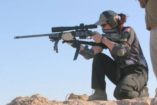 a woman in military fatigues kneels on the ground, aiming a scoped rifle with her right hand. she is wearing a tactical helmet, gloves, and elbow pads. she