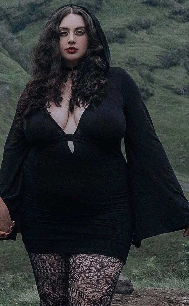 a woman with long, dark, curly hair and wearing a black hooded dress is standing in a field. the dress has a plunging neckline and long, flared sleeves. she is also wearing black lace tights and a choker. the background is a green hillside.