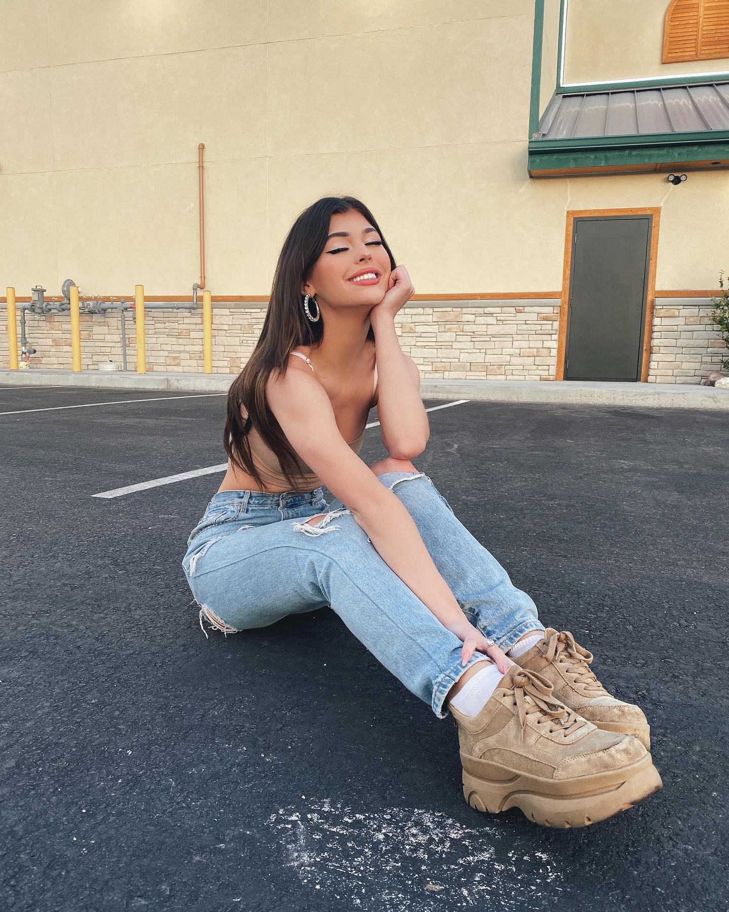 a young woman is sitting on an asphalt surface. she is wearing a lightcolored tank top, light blue ripped jeans, and tan platform shoes with white socks peeking out. she has long brown hair and is wearing hoop earrings. her eyes are closed and she is smiling with her head resting on her hand. in the background is a building with a light yellow exterior and a dark door. there are also metal fixtures on the wall of the building.