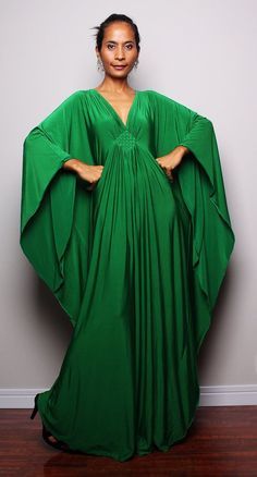 a woman stands indoors wearing a long, flowing green dress with wide sleeves, her hands on her hips. she is wearing jewelry and stands on a wooden floor against a light gray wall.