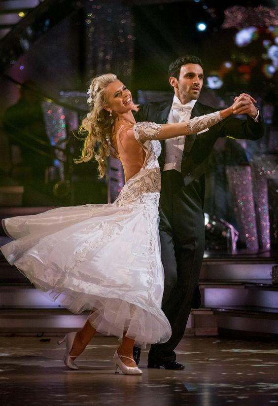 a woman in a white, glittering ballroom gown dances with a man in a black suit, likely performing a waltz on a stage under bright, sparkling spotlights.  the image captures the elegance and glamour of ballroom dancing.