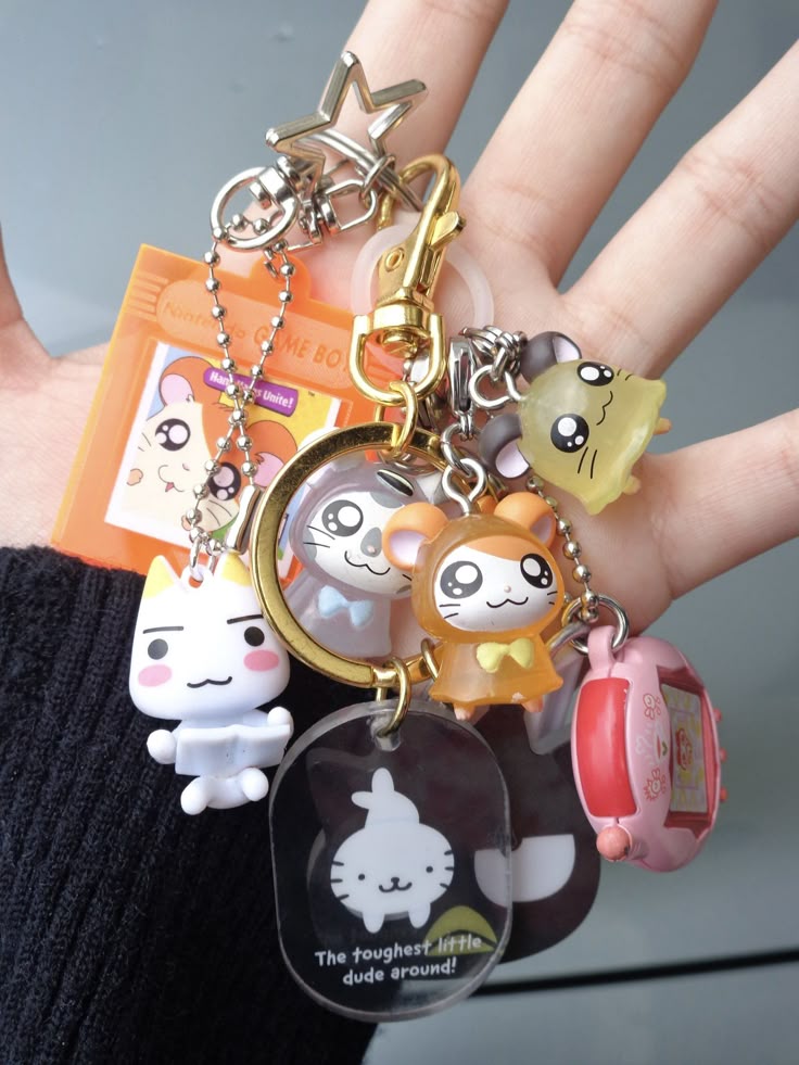 a hand holds a keychain with various charms, including animal figurines, a star, a game boy advance replica, and a miniature tamagotchi, all conveying a cute and playful aesthetic.