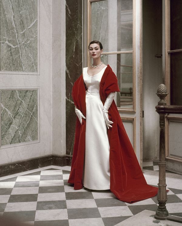 the image is a stunning black and white photograph, possibly by cecil beaton, showcasing a female model in a striking 1950s evening ensemble. she's wearing a pristine white satin gown, complemented by a dramatic floorlength crimson velvet cape. the cape drapes elegantly around her, creating a captivating contrast against the white dress.  the model is poised, her gloved hands gracefully resting at her waist.  she wears a simple necklace. the setting is a luxurious interior with a marble floor patterned in a black and white checkerboard design. a grand doorway and architectural details add to the sense of opulence.  the overall aesthetic is one of timeless elegance and high fashion.