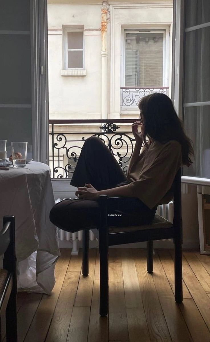 the image shows a woman sitting in a dark wooden chair next to a window overlooking a parisian street.  she appears relaxed and contemplative, almost silhouetted against the bright window.  she's wearing dark pants and a lightbeige top. the apartment has a sophisticated and slightly vintage feel, with a hardwood floor and an ornate balcony visible through the window. a table partially visible in the foreground suggests breakfast or a light meal. the overall mood is quiet and peaceful.
