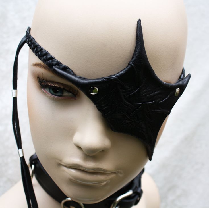 a closeup photo of a woman wearing a black leather eye patch.  the eye patch is decorated with a punkstyle design, and it is attached to a black leather headband. the woman is looking off to the side, and her expression is serious.