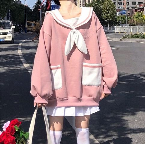 a woman is standing on a city street, wearing a pink sailor collar sweatshirt, a white skirt, and white socks. she is carrying a white tote bag and has her hair pulled back. the sweatshirt has a large white bow on the front and two large pockets. the image is taken from eye level and the lighting is bright and natural.