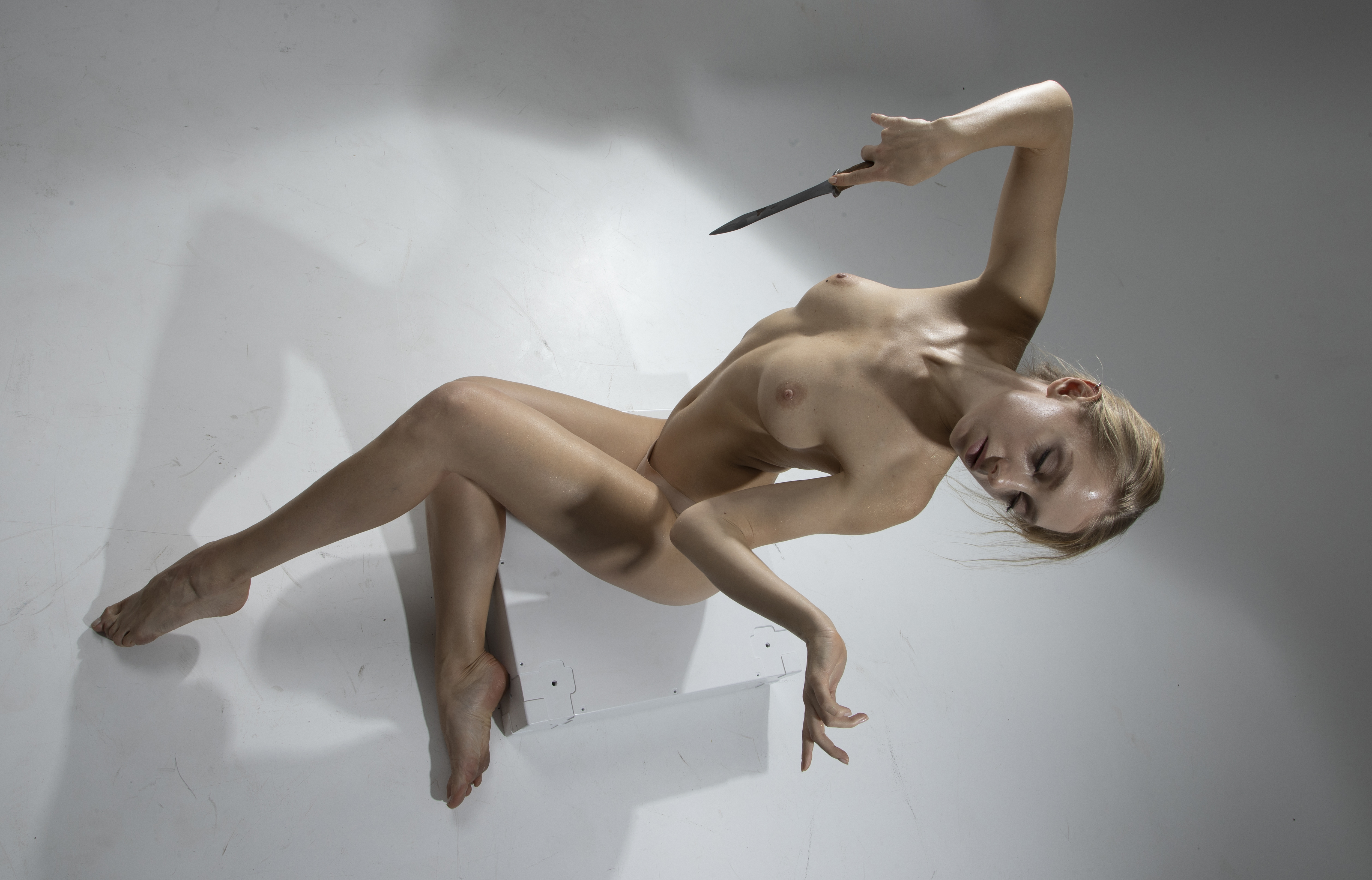 a nude woman with blonde hair is reclining in a studio setting, holding a knife above her head. her skin is pale and she is posed in a sensual manner.