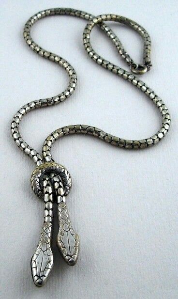 a closeup shot of a silver snake necklace on a white surface. the necklace features a chain made of small, interlocking circles. the pendant is shaped like two snake heads intertwined in a knot. the snake heads have detailed scales and facial features. the necklace is resting on a plain white background, which helps to highlight the details of the jewelry.