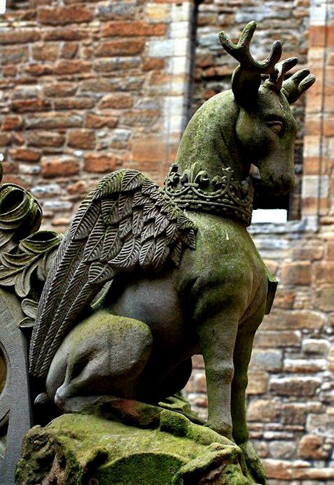 the image is a closeup of a weathered stone statue that depicts a mythical creature. it has the body of a deer, complete with a pair of small antlers. on its back, it has a set of feathered wings and a crown is around its neck. the statue is covered in moss, giving it a sense of age and history. it's positioned in front of a brick wall, which further emphasizes its historical setting. the architectural background suggests it may be part of a larger building or structure.