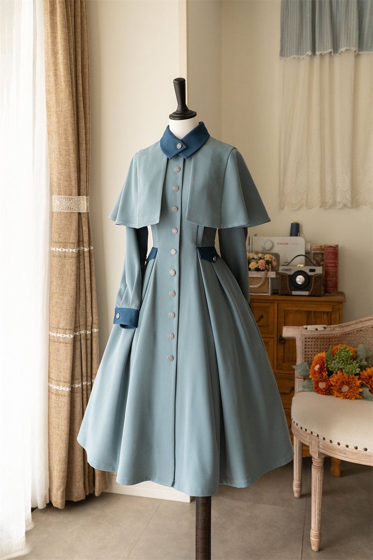 a blue vintagestyle dress with a cape is displayed on a mannequin in a naturally lit room. the dress has a full skirt, long sleeves, and a collared neckline. the cape drapes over the shoulders and fastens at the front.