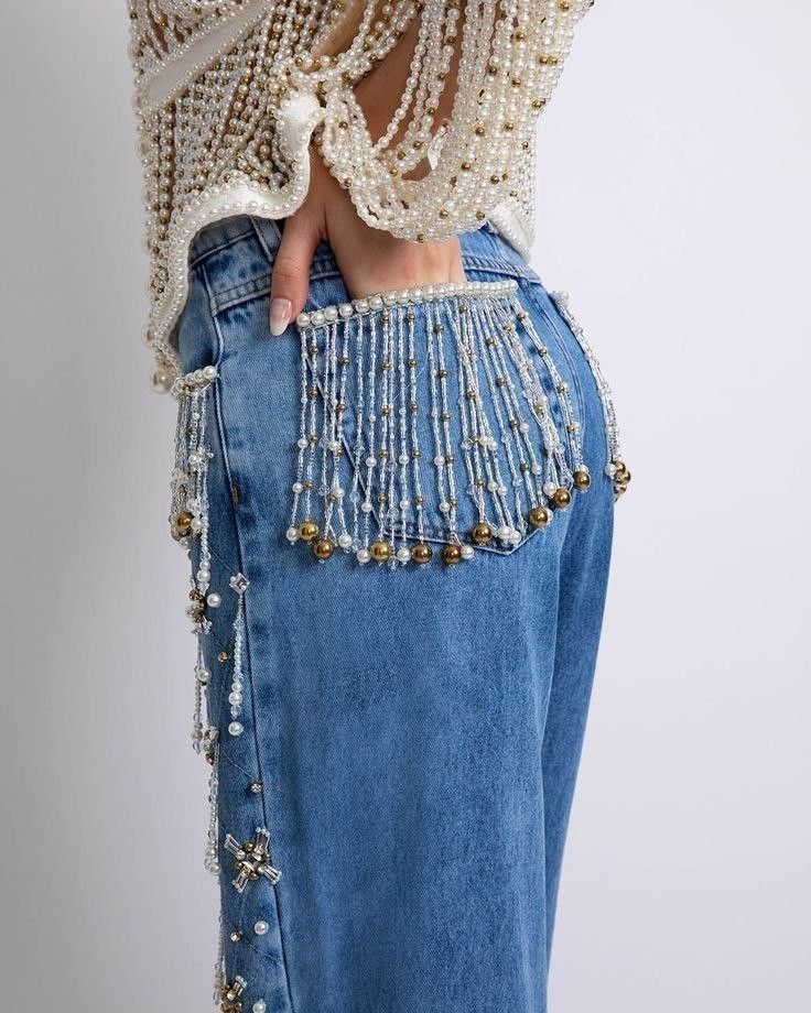 a closeup shot showcases blue denim jeans adorned with pearl and bead embellishments. the jeans feature a back pocket with a fringe made of white and gold beads. a woman's hand is seen with fingers tucked into the back pocket. the jeans are also decorated with beaded strands along the side seam. she is wearing a beaded top made of pearls and gold beads.