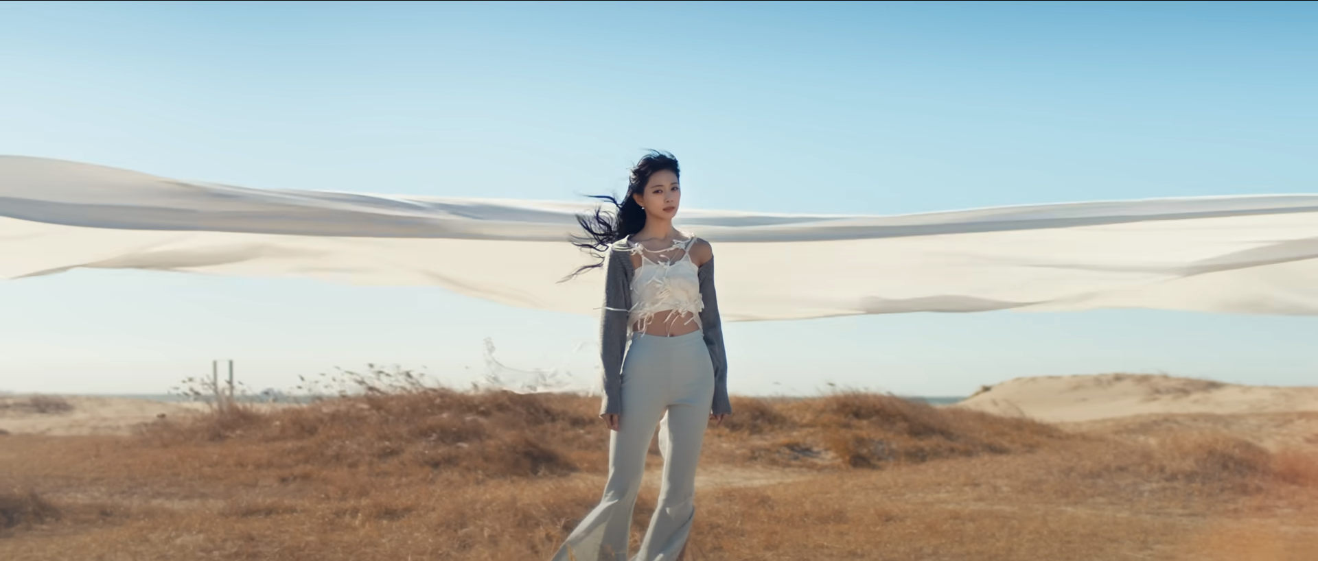 a woman stands on a beach with sand dunes in the background.  a large white sheet billows behind her.  she is wearing a white crop top, a grey cardigan, and grey pants.  the sky is blue and the sun is shining.