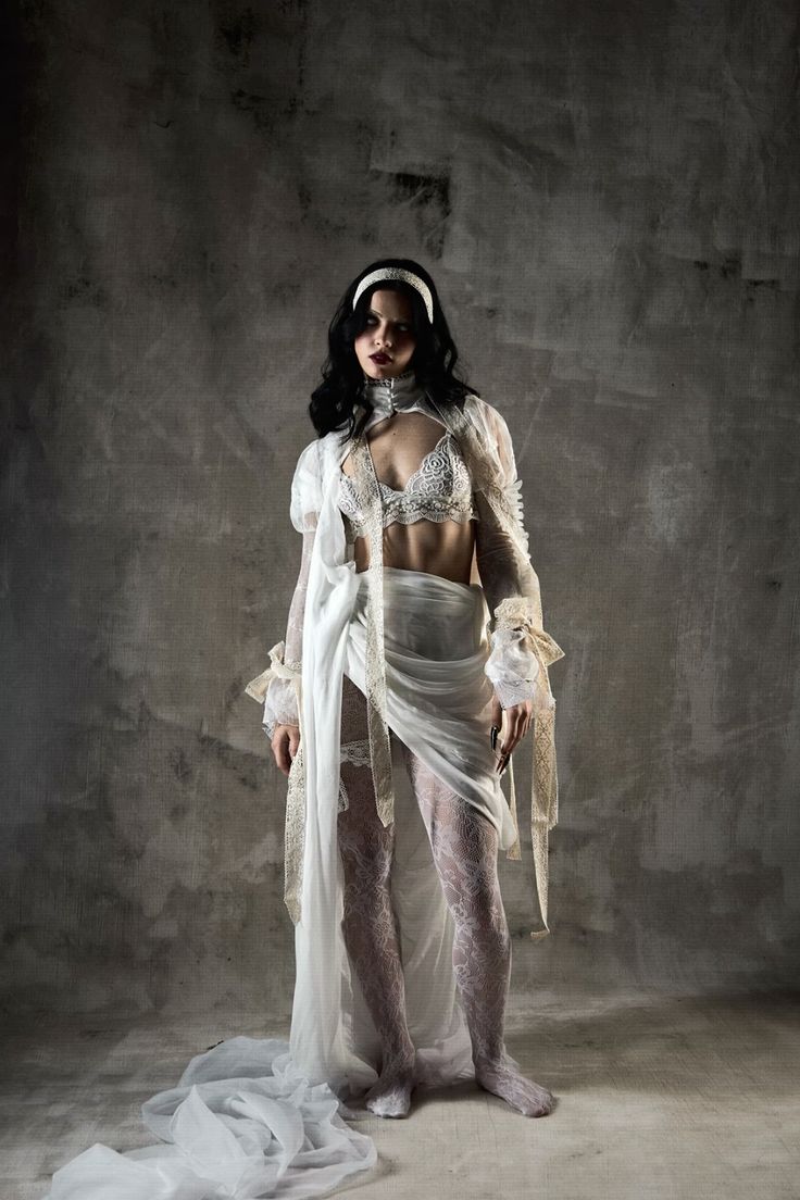a fulllength studio shot of a darkhaired woman posing in a white, tattered lace dress. the dress has a sheer quality and appears somewhat distressed or vintage in style. the model has a serious and slightly melancholic expression, and the overall mood is dark and artistic, with strong contrast in the lighting. the background is a textured grey wall, enhancing the image's dramatic atmosphere. the style could be described as gothic or alternative bridal. the image suggests a concept or artistic vision, rather than a simple fashion photograph.