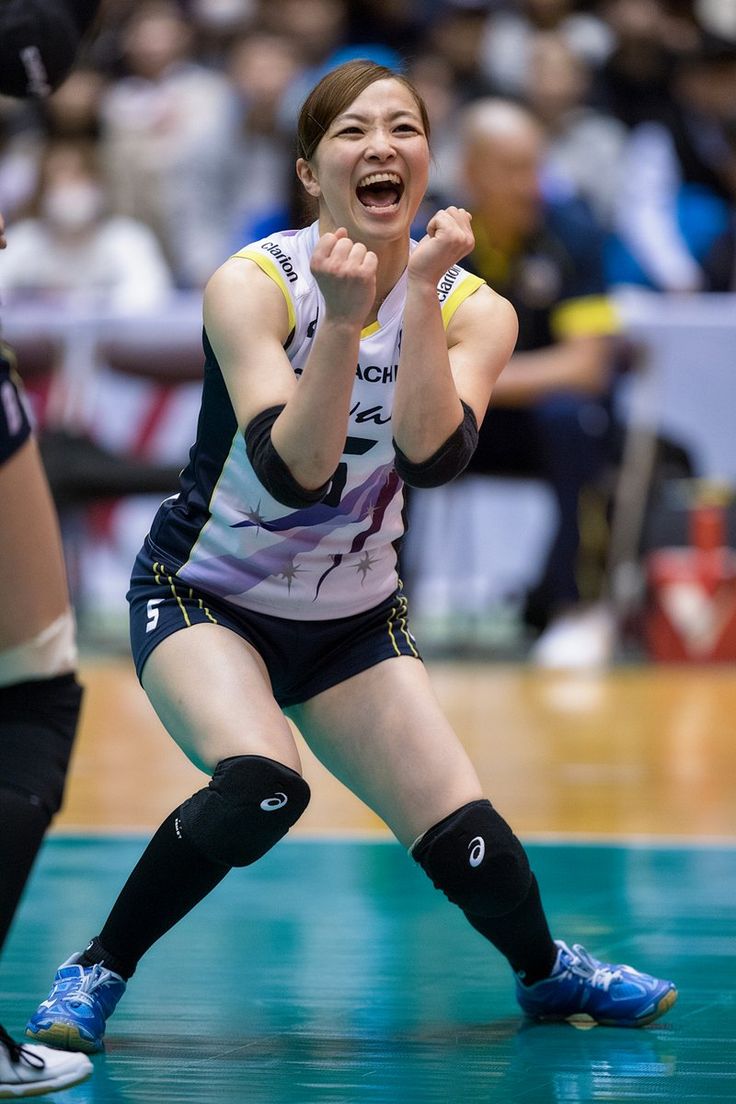 a female volleyball player in a blue, white, and yellow uniform celebrates a point with a triumphant yell. she's on an indoor court, captured in midair with her arms raised in victory. the perspective is from slightly below, emphasizing her energy.