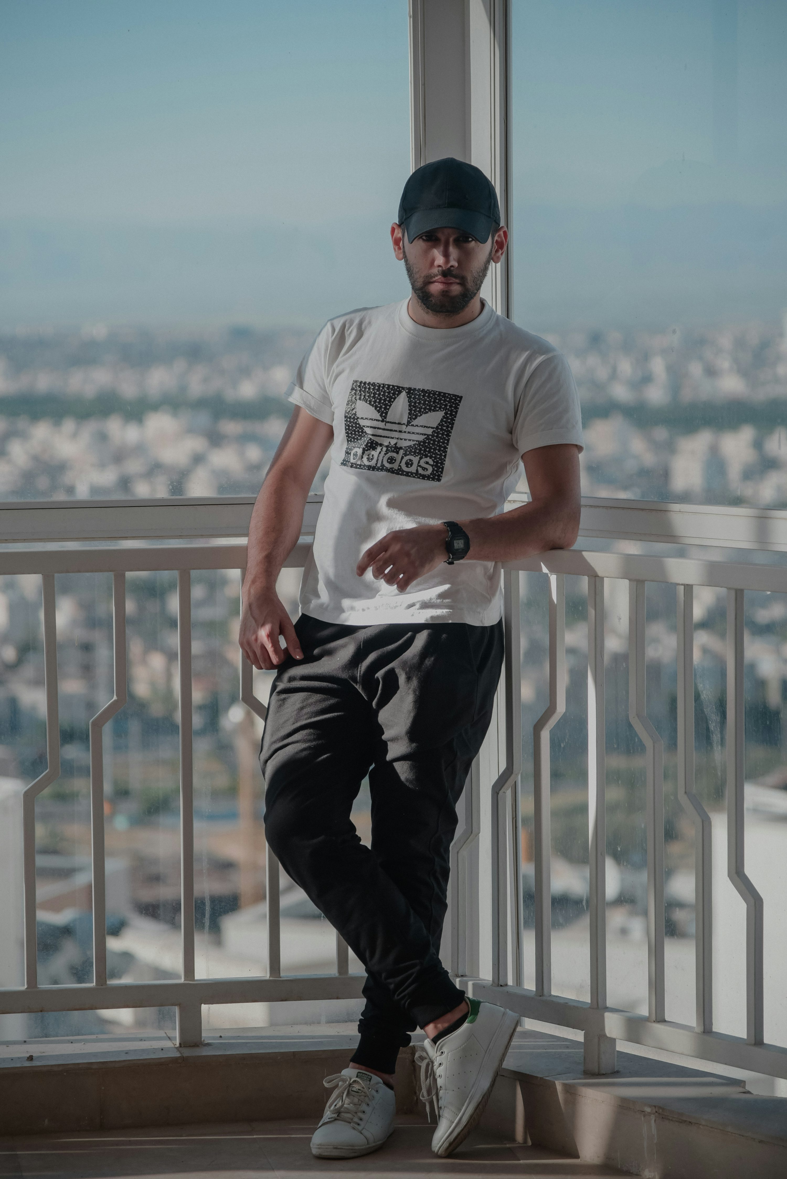 a young man is standing on a balcony overlooking a city. he is wearing a white adidas tshirt, black joggers, white sneakers, a black baseball cap, and a watch. he is leaning against the railing and looking at the camera with a serious expression. the photo is taken outdoors on a sunny day.