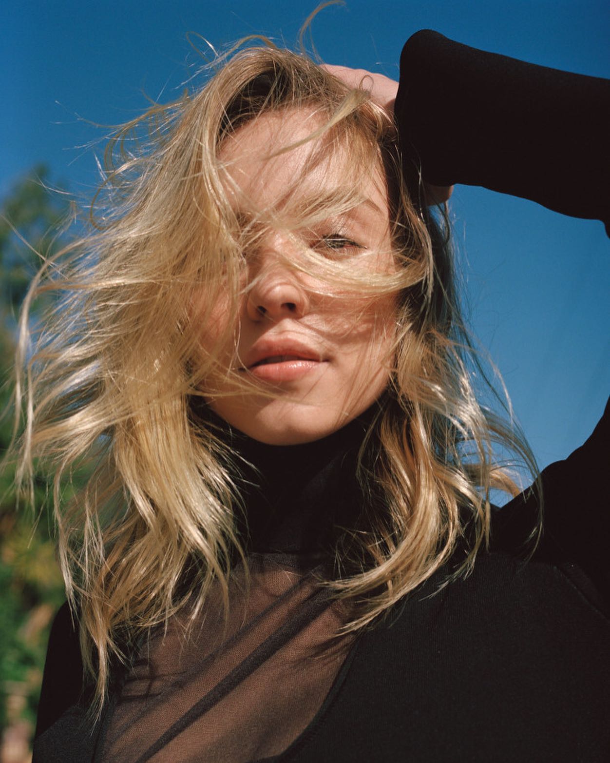 a closeup portrait of a blonde woman with windblown hair. she is outdoors on a sunny day with a bright blue sky in the background.  she is wearing a black sheer turtleneck top layered over a black turtleneck. the woman is looking slightly downwards, and her hair partially obscures her face. the overall mood is casual and natural, highlighting her beauty and the feel of the wind. the image appears to be a fashion or editorial style photograph.