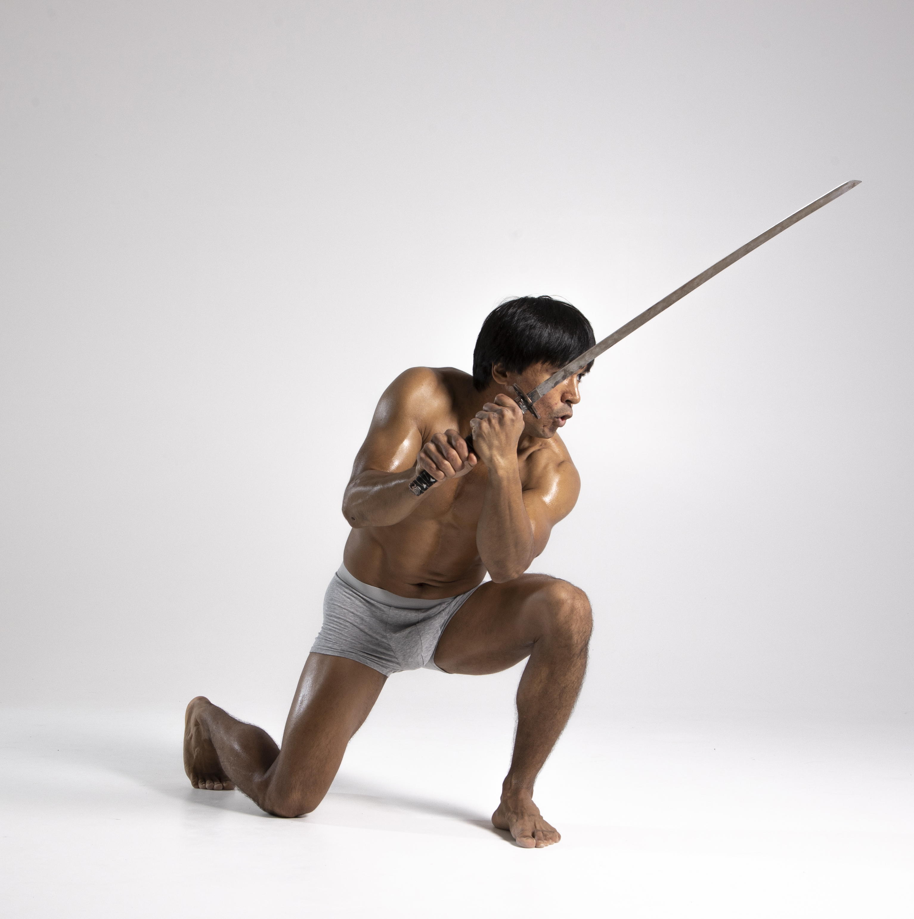 a shirtless asian man in gray underwear is kneeling with one leg on the ground and the other bent at a 90degree angle. he is holding a sword up in a defensive posture.