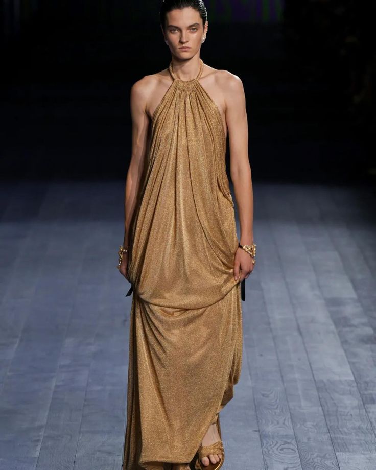 a model walks the runway in a long, gold halter dress. the dress has a sparkly, metallic finish and is sleeveless. the model is wearing gold jewelry and her hair is pulled back.  the runway is made of wood planks and the background is blurred.  the model is walking toward the camera.