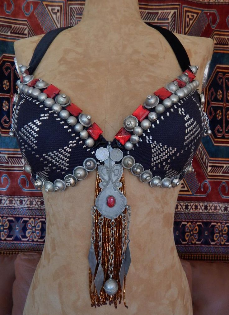 a closeup shot of a traditional belly dancing bra. the bra is made of black fabric with intricate silver and red embellishments. the bra is worn on a mannequin in front of a colorful rug. the bra is a beautiful example of the craftsmanship and artistry that goes into traditional belly dancing costumes. it's a very unique and decorative piece of clothing.
