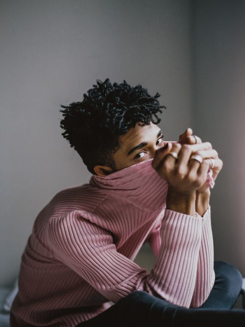 a young black man is sitting on a white surface. he is wearing a pink sweater, his hands are covering his face with the sweater. he has black curly hair and he looks serious. the background is a grey wall.