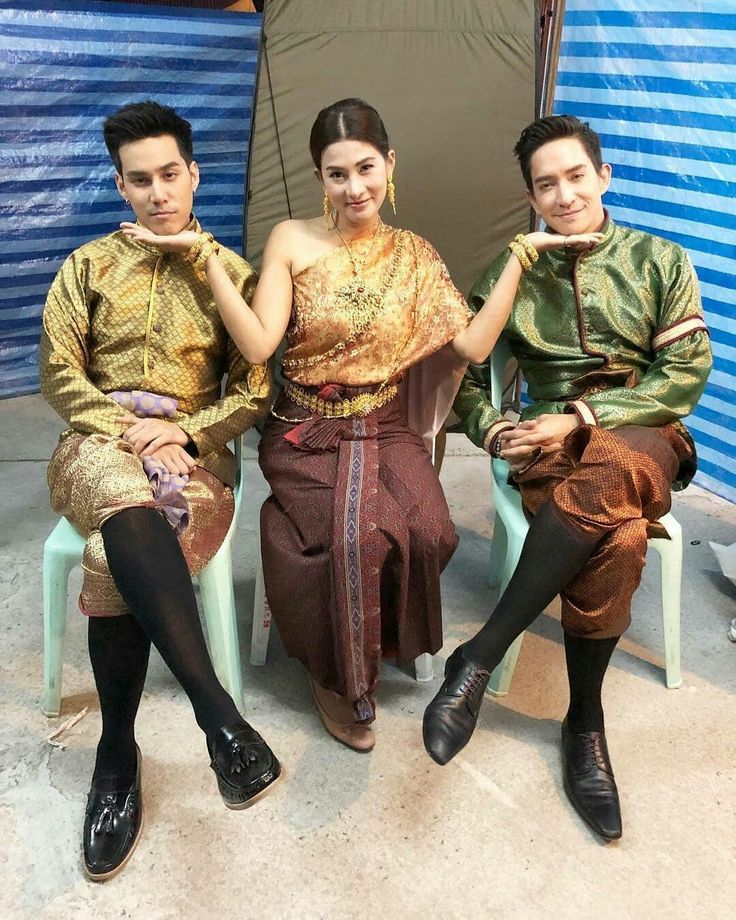 three people, two men and one woman, are sitting and smiling at the camera in traditional thai clothing. the woman is wearing a gold, brown, and maroon outfit and the men are wearing gold and green outfits. they are sitting in a room that appears to be indoors.
