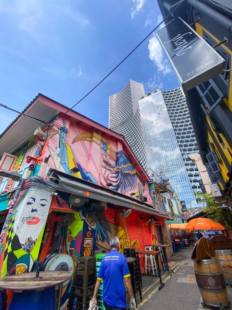 a vibrant and colorful alleyway in singapore, filled with street art and graffiti on the buildings. a man in a blue shirt walks through the alley, which is lined with outdoor seating for a cafe or bar. in the background, modern skyscrapers tower above the traditional shophouses. the scene is sunny and lively.