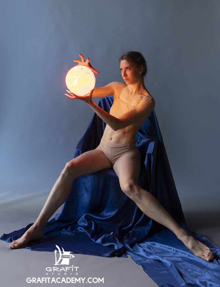 a woman in a dance pose, seated on flowing blue fabric, holds a glowing orb in her hands. the background is a cool blue, contrasting with the warm light emanating from the orb. the scene appears to be in a studio setting.