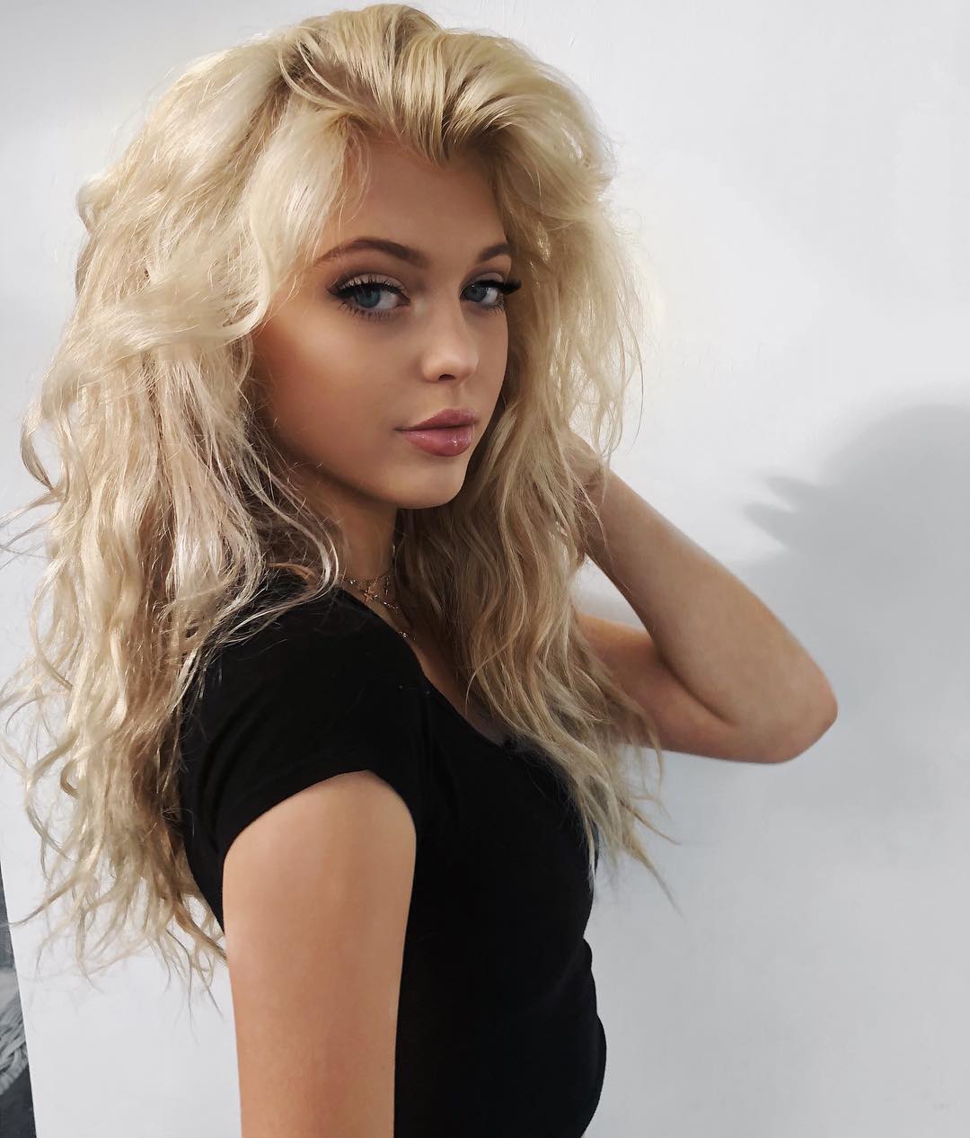 a young woman with blonde, curly hair and blue eyes is posing in front of a white wall. she is wearing a black shirt and looking directly at the camera. her hair is styled in loose waves and she has a fair skin complexion. she is wearing lipstick.
