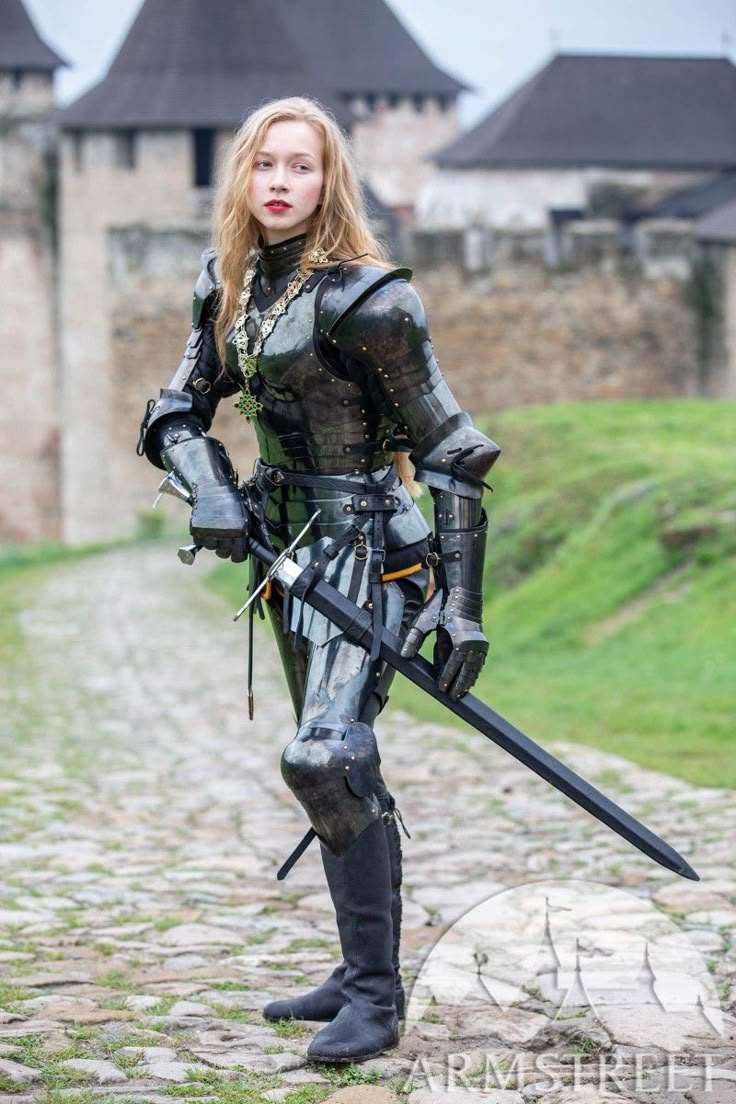 a woman with blonde hair is standing in full plate armor and holding a sword. she is standing on a stone path in front of a castle. the armor is dark and shiny, and she is wearing black boots. she also has a chain necklace. the setting suggests a medieval or historical fantasy theme. the brand "armstreet" is visible in the bottom right corner.