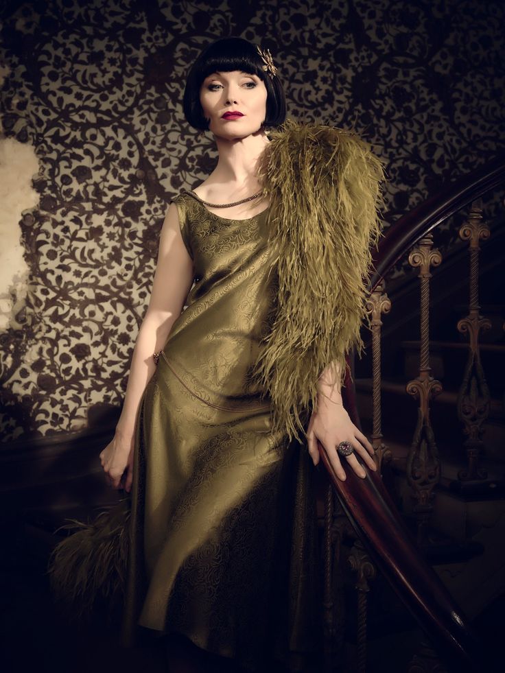 a woman in a vintage dress stands on a staircase, holding the railing. she wears a stylish hat with a feather boa. the woman is in a glamorous pose and looks very elegant.