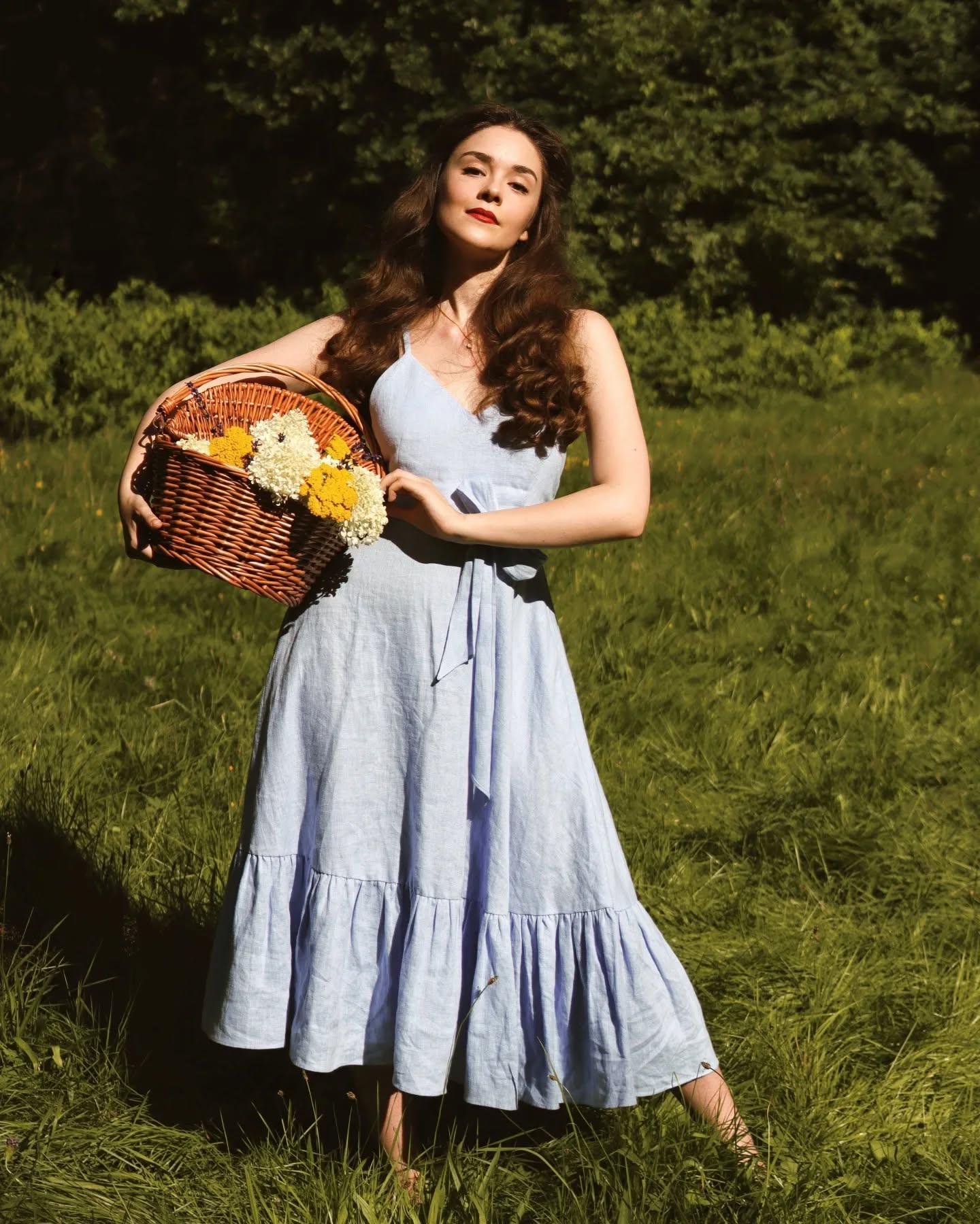 a beautiful young woman with long brown hair is standing in a sunny green field. she is wearing a light blue linen maxi dress with ruffles and is carrying a wicker picnic basket filled with wildflowers. she is barefoot and looking directly at the camera with a happy expression. the image is bright and cheerful, evoking a feeling of summer and carefree joy.