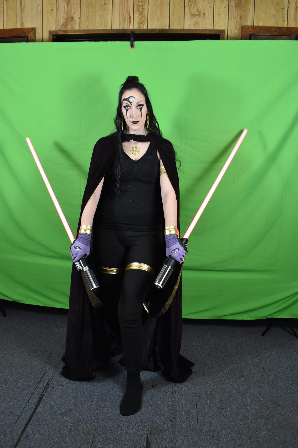a fullbody shot of a woman cosplaying as a sith character from star wars. she's wearing a black costume with a cape, golden accents, and purple gloves.  she has intricate face paint and is holding two lightsabers. the background is a green screen. the overall impression is dark and powerful.