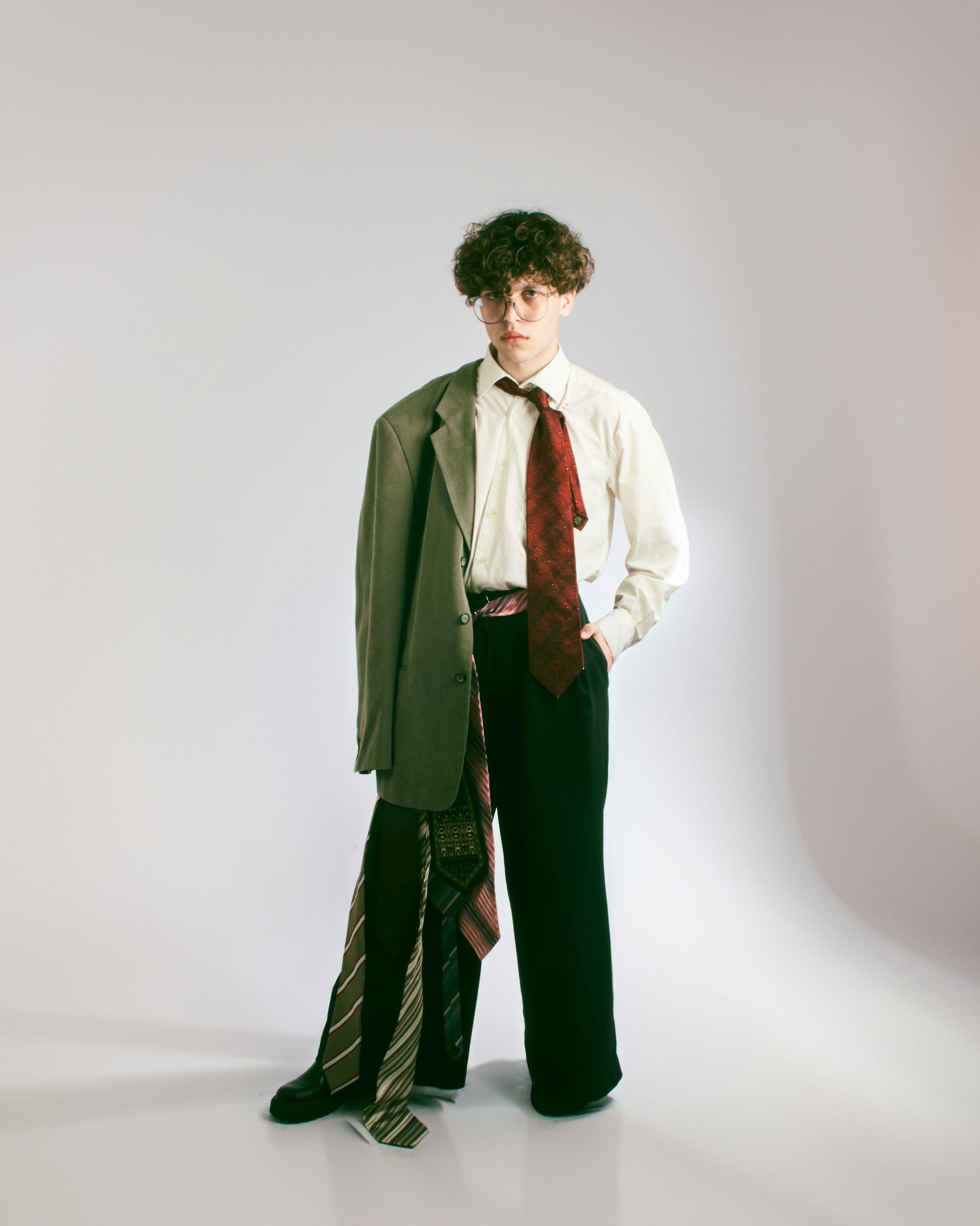a young man with curly brown hair and glasses is standing against a light gray background. he's wearing a white shirt, dark trousers, and a green jacket draped over his shoulder.  several ties are casually draped around his legs and hanging from the jacket. the overall style is a blend of sophisticated and youthful, with a slightly unconventional twist. the image is a studio portrait, sharply focused and welllit.
