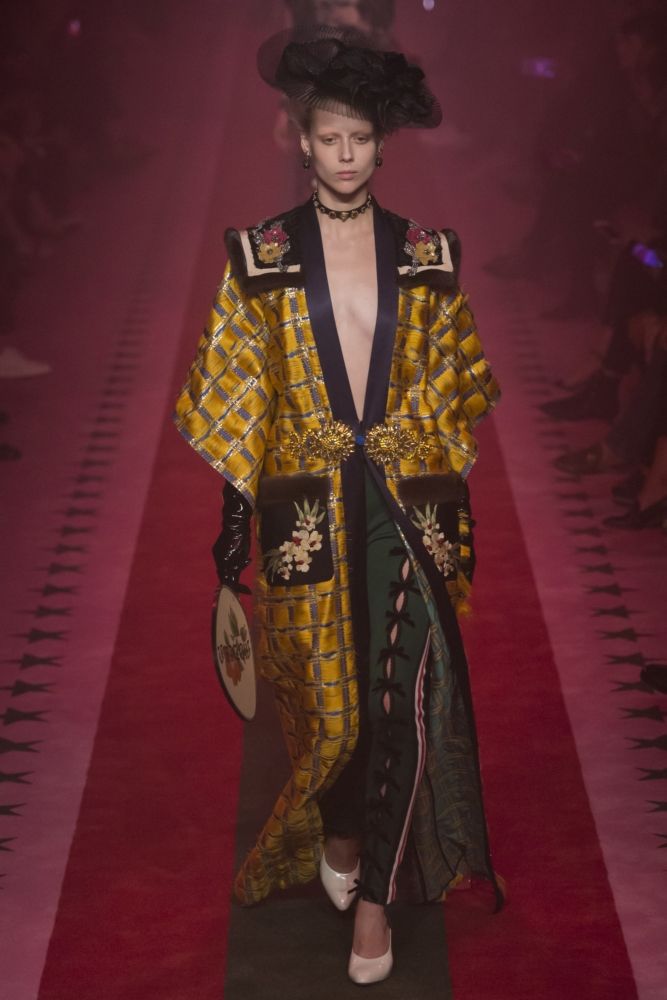 a model walks down a runway during a fashion show, wearing a long, gold and black plaid coat with embroidered details. the coat is worn over a dark green and black dress. she also wears a black hat, black gloves, and white shoes. the runway is carpeted in red, and the background is dimly lit in shades of purple and pink.