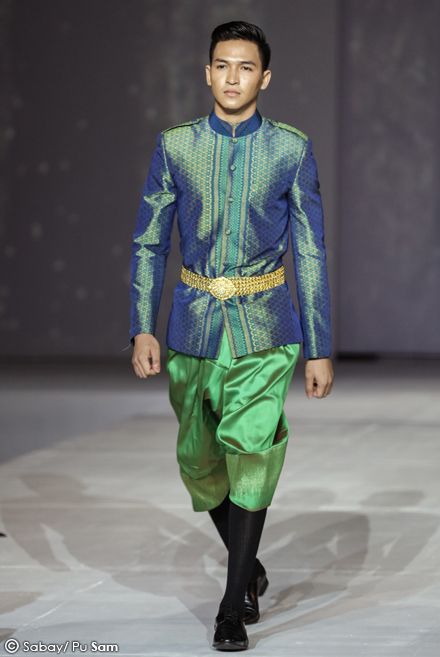 a male model walks the runway in a traditional thai outfit. he is wearing a blue silk shirt with gold trim, green silk pants, and a gold belt. he is also wearing black shoes. the model is looking straight ahead and smiling.