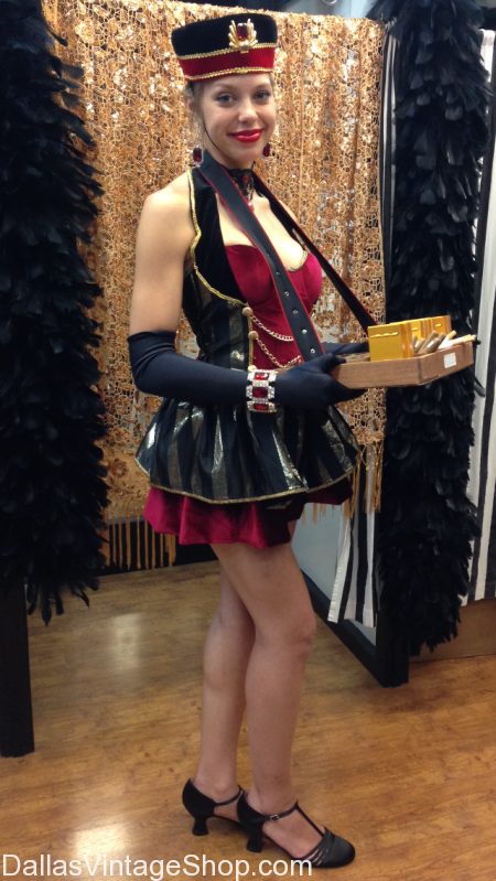 a fulllength shot of a woman dressed in a vintage burlesque showgirl costume.  she is wearing a black and gold dress with red accents, a matching hat, long black gloves, and black shoes.  she holds a tray with what appears to be cigars. the background features a gold and black striped backdrop and black feather boas. the photo is taken in what appears to be a vintage shop.  the watermark "dallasvintageshop.com" is present at the bottom.