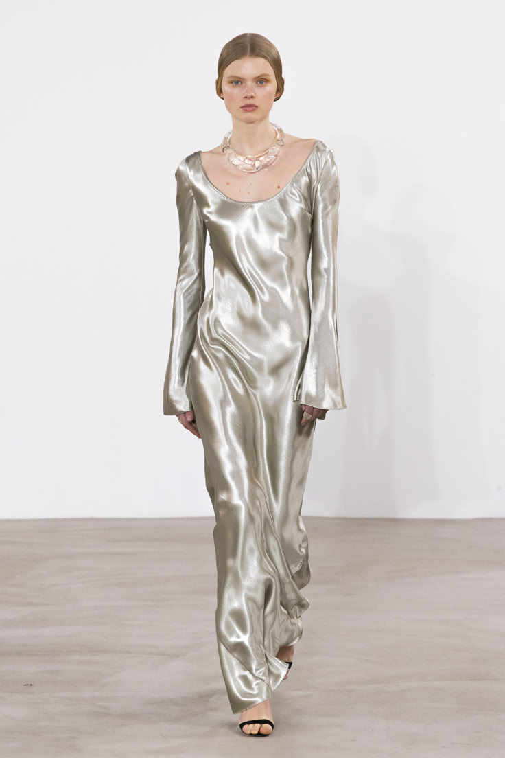 a woman is walking down a runway wearing a long silver satin dress with long, bellshaped sleeves, a clear necklace, and black sandals.