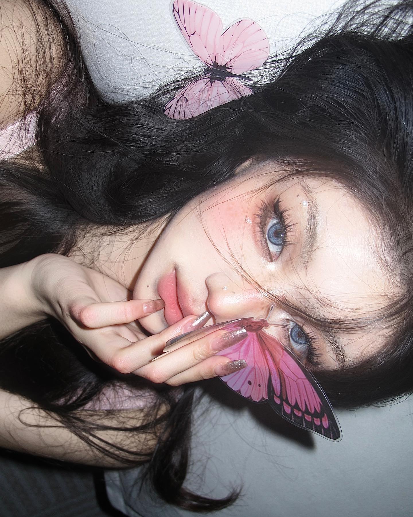 a closeup selfie of a young woman with long dark hair, butterfly decorations, and elaborate makeup. she is lying down and looking directly at the camera. the image has a soft, dreamy quality.
