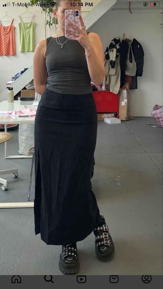 a woman is taking a mirror selfie inside a brightly lit studio. she's wearing a fitted pinstriped black maxi skirt with a thighhigh slit, a dark green tank top, and chunky black platform boots with silver buckles. her outfit is simple, yet stylish. she's holding up her phone to take the picture. in the background, we can see a clothing rack with colorful garments and some storage boxes.