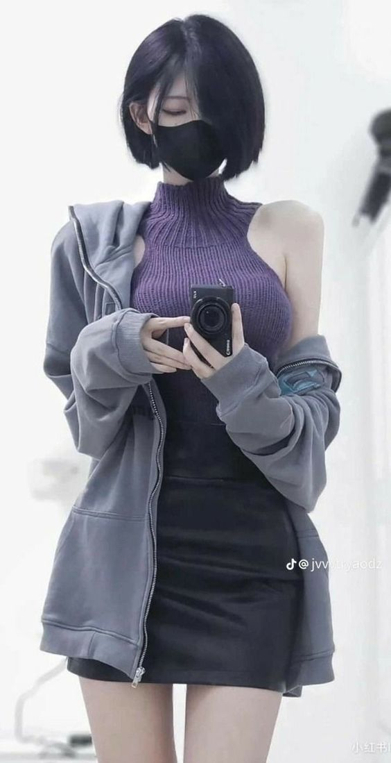 a young woman takes a mirror selfie. she is wearing a black face mask, a purple turtleneck sleeveless top, a gray zipup hoodie, and a black leather skirt. she has short black hair and is posing with one hand in her hoodie pocket and the other holding her phone up to take the picture. the background is white and out of focus.