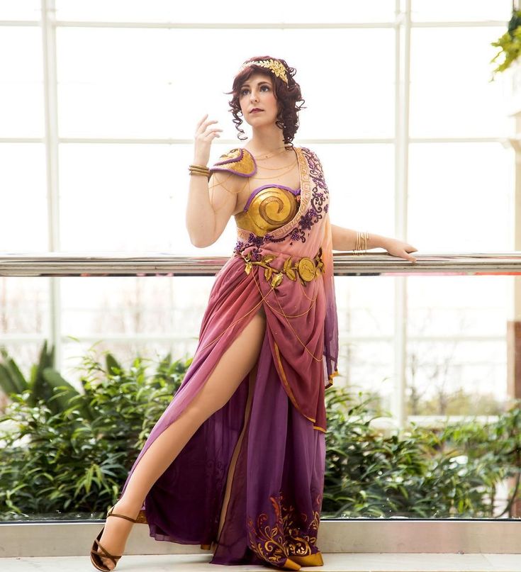 a woman is posing for a photo while wearing a cosplay outfit of a greek goddess. she is wearing a long, flowing purple and gold dress with a high slit, gold sandals, gold bracelets, a gold headpiece, and gold shoulder armor. she has curly brown hair and is standing in front of a window with plants behind her. she is leaning on a railing.