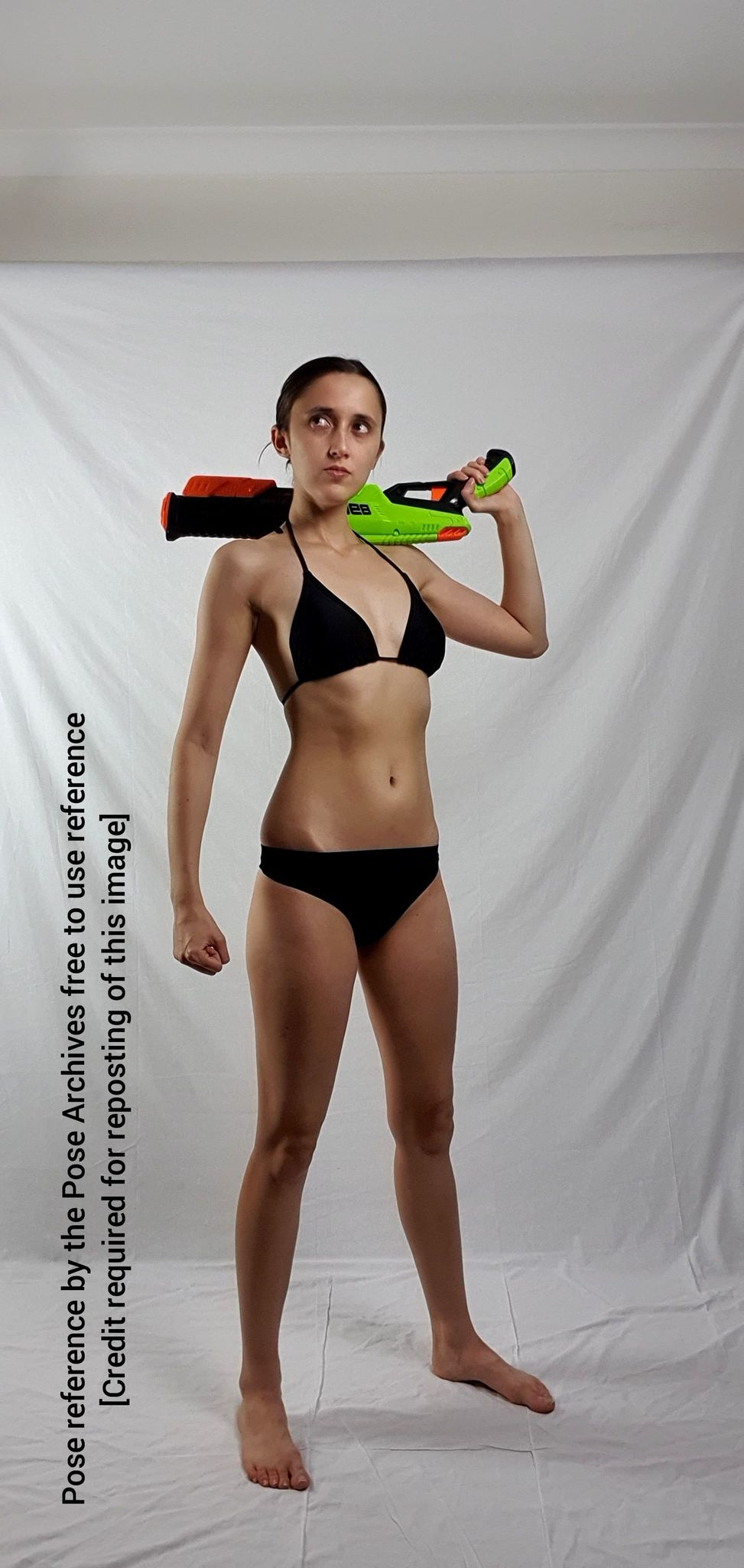 a young woman in a black bikini is standing against a white backdrop. she is holding a green and orange toy gun over her shoulder.