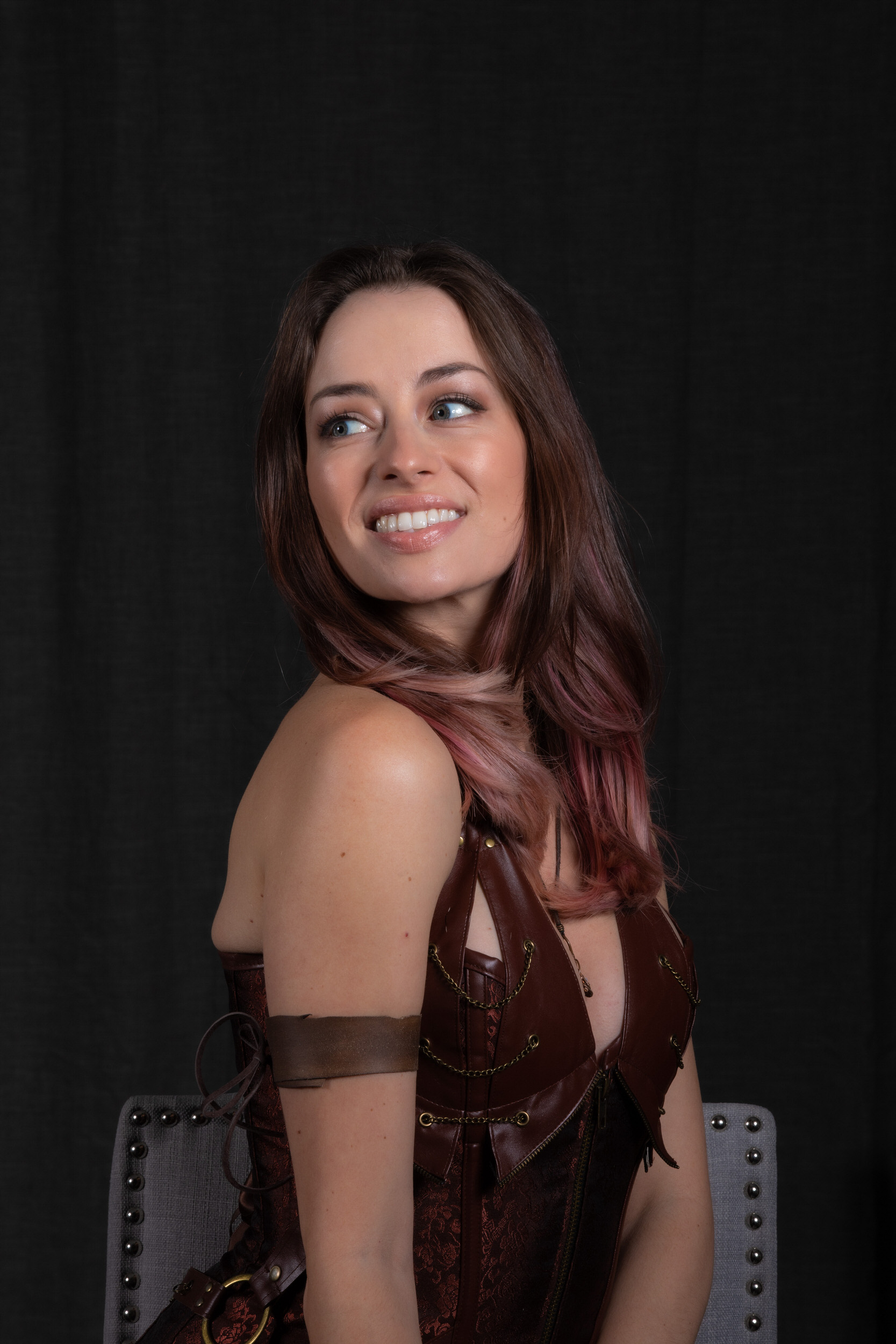 a brunette woman with pink highlights in her hair sits facing slightly away from the camera, her head turned to look over her left shoulder with a pleasant smile.  she wears a brown leather and brocade corset with metal accents and a brown leather bracer on her right arm. the background is a plain black backdrop and she is posed against a grey fabric chair back with metal studs.  the image is lit with studio lighting.