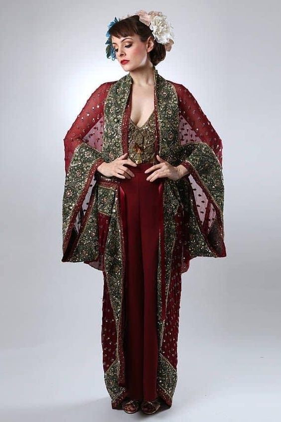 a woman stands in a studio wearing a dark red outfit, which includes a vneck top, high waisted straight leg pants and a long, red, sheer kimono with dark green and gold embroidery. she has her hands on her hips and a large flower crown on her head.