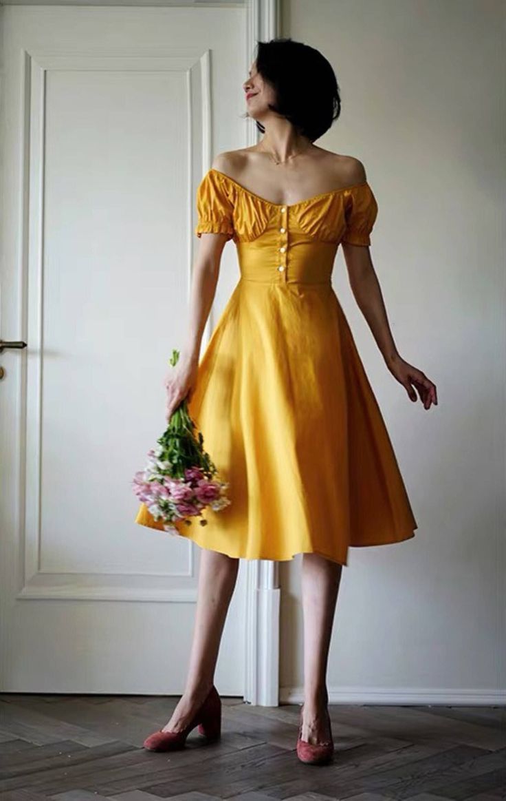 a young woman stands in front of a white door in a yellow dress. she is holding a floral bouquet and is wearing red shoes. the woman is posing for the camera and has a pretty smile. she has short black hair and a stylish dress. the wood floor in the background adds a warm element to the image.