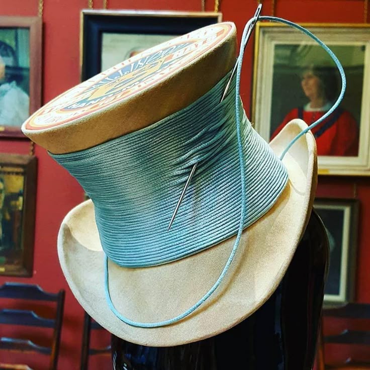 a closeup shot of a whimsical hat shaped like a giant spool of thread, complete with a needle and blue thread wound around the spool. the hat is tan with a large, colorful, circular top resembling a vintage thread spool label. a needle is inserted into the blue thread wrapped around the middle. the hat sits on a mannequin head. in the background, a red wall is adorned with framed artwork and paintings. the scene is captured in a still life style with elements of antique and vintage craftsmanship.