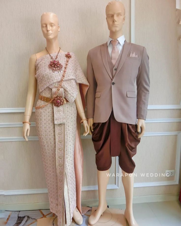 a thai bride and groom in traditional attire. the bride is wearing a long, pink and gold dress with intricate beading and a flower on her head. the groom is wearing a brown suit with a vest and a tie. the background is a light beige wall. the bride and groom are both smiling and look happy. they are standing side by side with the bride on the left and the groom on the right. the bride is holding a bouquet of flowers. the groom is holding a white dove.
