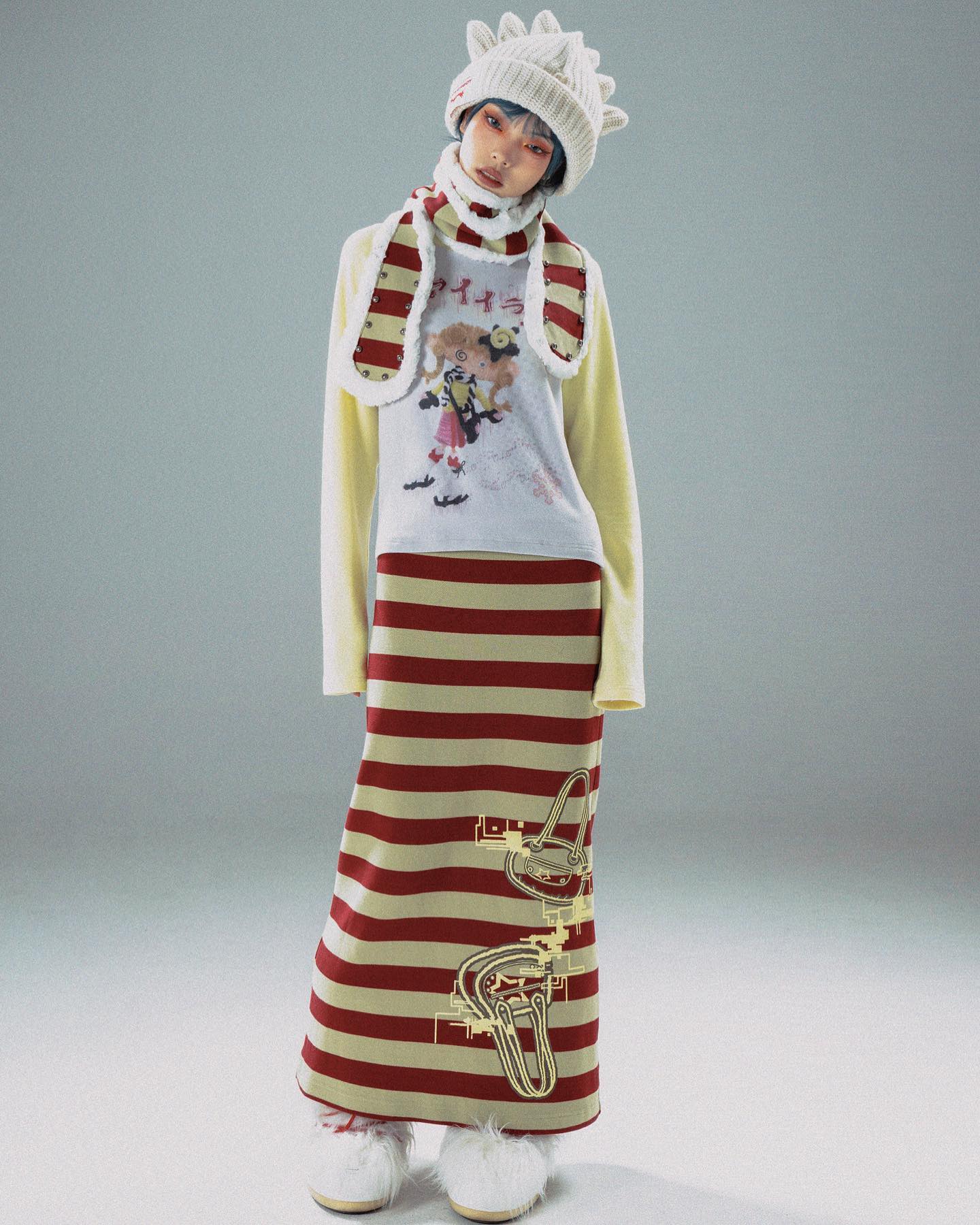 the image showcases a fashion model wearing a distinctive ensemble. she's dressed in a longsleeved, light yellow sweater featuring a whimsical animestyle graphic print. this is paired with a long, horizontally striped maxi skirt in red and beige featuring additional graphic elements. the model also sports a creamcolored, textured knit beanie with a slightly spiky design, and a matching scarf with red stripes. her footwear consists of fluffy, creamcolored boots. the overall style is unique, blending elements of streetwear, anime, and quirky design. the setting is a simple lightgrey studio backdrop, which places all the emphasis on the clothing itself.