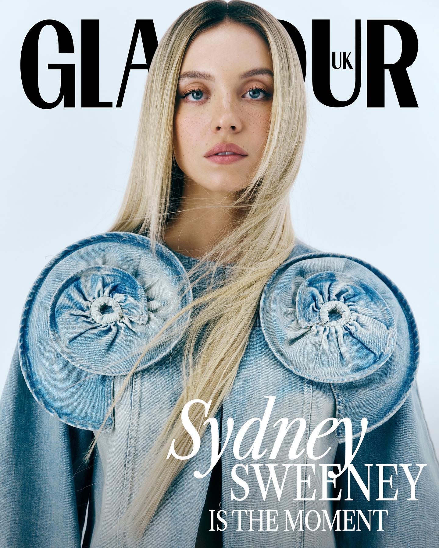 the image is a magazine cover featuring actress sydney sweeney for glamour uk. she is shown in a closeup portrait, with long blonde hair flowing down. sweeney is wearing a light wash denim jacket adorned with large, decorative denim rosettes on the shoulders. the background is a simple, light color. the glamour uk logo is prominently displayed at the top, and text underneath reads "sydney sweeney is the moment." the overall aesthetic is fashionforward and sophisticated.